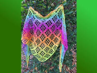 Gehäkeltes Dreieckstuch mit buntem Farbverlauf und Rautenmuster, aufgehängt vor grünen Blättern