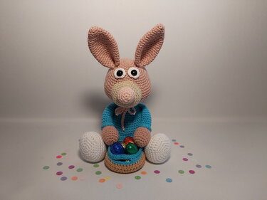 Sitzender Amigurumi-Osterhase mit blauem Pullover und Häkelkörbchen mit bunten Eiern.