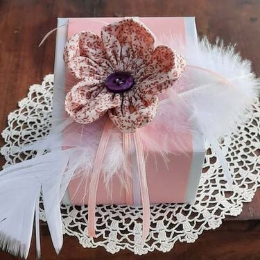 Detailaufnahme: Stoffblume mit lila Knopf, Satinbändern und weißen Federn auf rosa Geschenkbox
