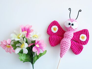 Pinker gehäkelter Schmetterling auf Holzstab neben rosa-weißen Kunstblumen