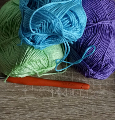 Mint green, aqua, and purple yarn skeins with an orange crochet hook on a wooden surface
