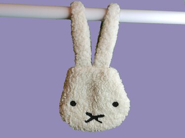 Flauschige weiße Plüschtasche in Kaninchenform mit langen Ohren, schwarzen Augen und X-förmigem Mund, hängend an einer Stange vor lila Hintergrund