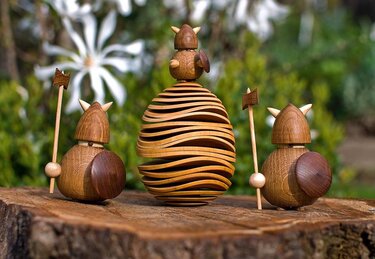 Holzfiguren mit runden Körpern, Helmen und Schilden neben einer wellenförmig geschichteten Holzeierskulptur
