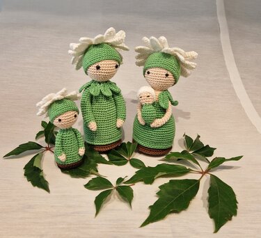 Drei gehäkelte Blumenpuppen in Grün mit weißen Blütenhüten, eine hält ein Baby; Laub als Dekoration