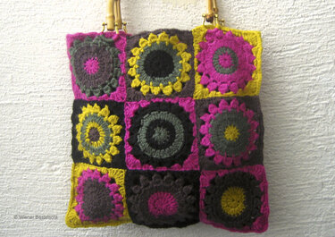 Gehäkelte Henkeltasche aus neun Granny Squares in Fuchsia, Gelb und Dunkelgrau mit Bambusgriffen vor heller Wand