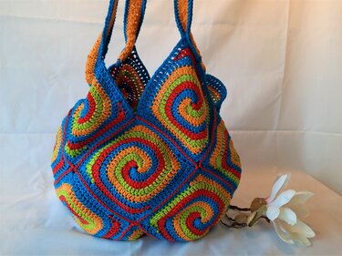 Handgehäkelter Shopper aus bunten Granny Squares mit Spiralmuster neben einer weißen Blüte