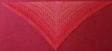 Rotes bis orangefarbenes gehäkeltes Dreieckstuch mit Lochmuster, flach auf rotem Hintergrund gelegt.