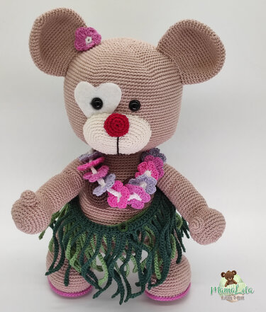 Gehäkelter Amigurumi-Teddybär in Beige mit grünem Hula-Rock, rosa-lila Blumenkette, weißem herzförmigen Augen-Patch und pinker Blüte am Ohr vor hellem Hintergrund.