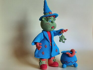 Gehäkelte grüne Hexe mit blauem Hut und Mantel hält einen Frosch, daneben ein blauer Kessel.