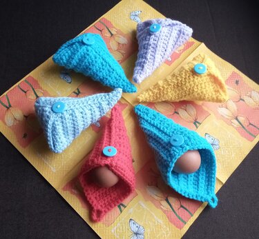 Sechs bunte handgehäkelte Eierwärmer mit Knöpfen auf gelber Serviette, teils mit braunen Eiern