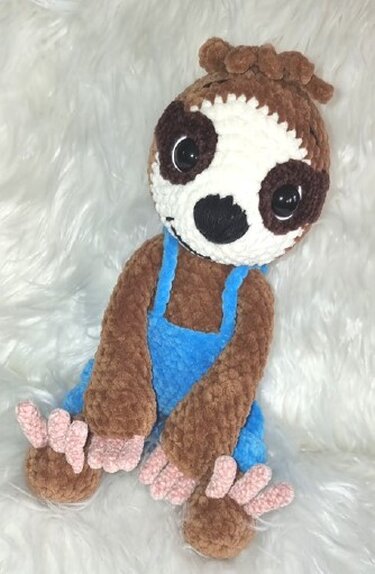 Gehäkeltes Faultier-Amigurumi in Braun und Weiß mit blauer Latzhose und rosa Krallen auf weißem Fell