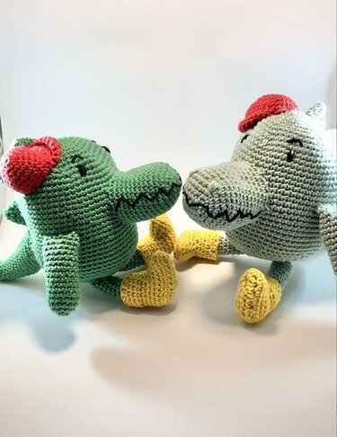 Zwei Amigurumi-Krokodile, grün und hellgrau, mit roten Mützen und gelben Füßen auf weißem Hintergrund.