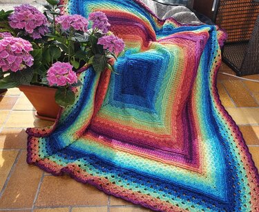 Gehäkelte quadratische Decke mit Regenbogenfarbverlauf und Spitzenrand, drapiert neben einem Topf mit rosa Hortensien auf gefliesten Boden