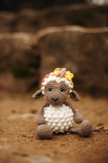 Gehäkeltes Schaf-Amigurumi mit runder Brille und Blumenmütze auf sandigem Untergrund.