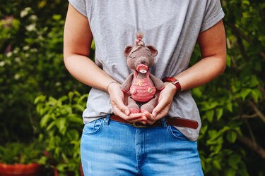 Hand hält gehäkelten Amigurumi-Teddybär in rosa Latzhose mit Schnuller vor grünem Hintergrund.