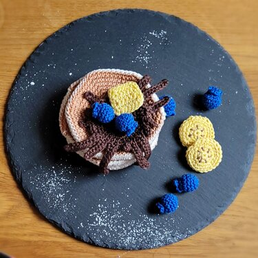 Mehrteiliges Häkelspielset mit Pfannkuchen, 'Butter', Sirup, Blaubeeren und Bananenscheiben auf runder Schieferplatte