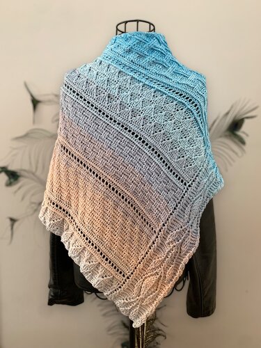 Blue-to-peach ombre crocheted triangle shawl with lace motifs, textured stitches, eyelet rows, and ruffled edge on a mannequin.