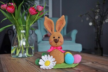 Gehäkelter brauner Amigurumi-Hase mit rosa Schleife, bunten Ostereiern und Gänseblümchen auf Holztisch