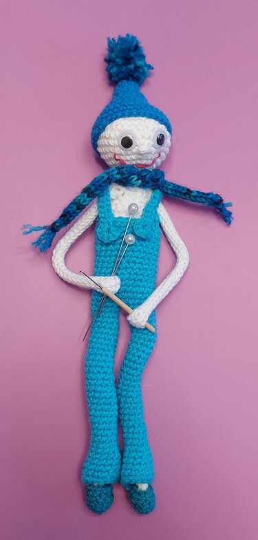 Gehäkelte Amigurumi-Puppe in blauem Outfit mit Mütze, Schal und Nadeln