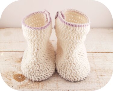 Ein Paar cremefarbene, gehäkelte Babystiefelchen mit lila Rand und Knöpfen auf hellem Holzhintergrund.