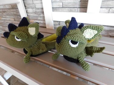 Zwei grüne gehäkelte Drachen-Hausschuhe mit dunkelblauen Stacheln und weißen Augen auf einer Holzbank.