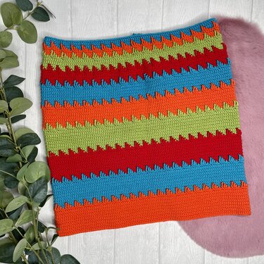 Gehäkelter Rock mit horizontalen Streifen in Rot, Blau, Orange und Hellgrün, auf hellem Holzboden liegend mit grünen Blättern links und rosafarbenem Fell rechts.