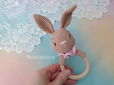 Beige crocheted bunny head rattle with a pink ruffle attached to a wooden teething ring, held by a hand.
