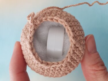 Hand holding crocheted rattle cover revealing fiber stuffing and a plastic inner ring