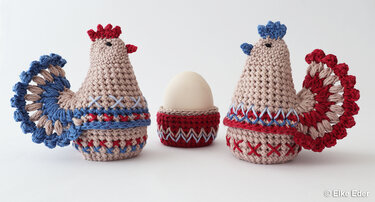 Two crocheted chicken egg cozies (rooster and hen) with a matching crocheted egg cup holding a white egg, in beige, red, and blue.