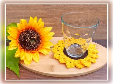 Clear glass resting on a yellow and brown crocheted sunflower coaster next to a sunflower on a wooden board.