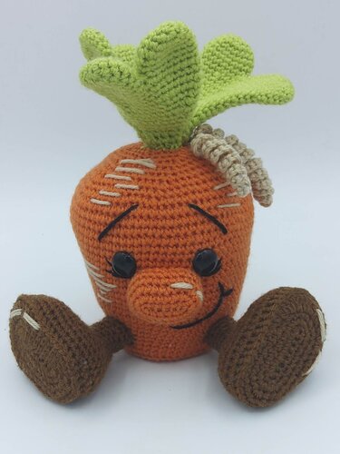 Gehäkelte Amigurumi-Karotte in Orange mit grünen Blättern, schwarzen Knopfaugen und braunen Füßen vor hellem Hintergrund.