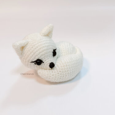 White crocheted fox amigurumi curled into a ring with black stitched eyes and nose