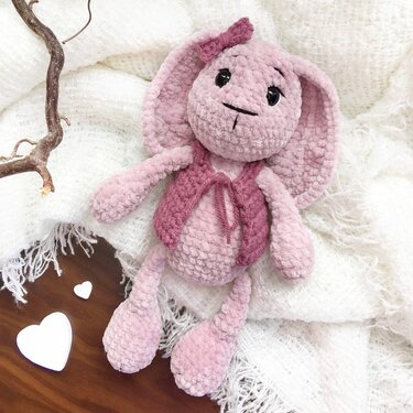 Rosa gehäkelter Amigurumi‑Hase mit Weste und Schleife auf weißer Decke, Ast und zwei Holzherzen daneben