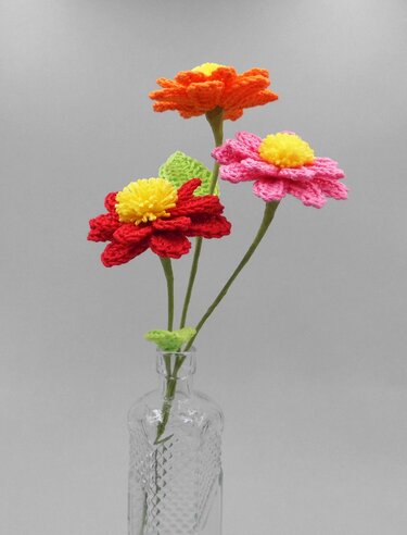 Three crocheted flowers (red, pink, orange) with yellow centers in a clear glass bottle vase