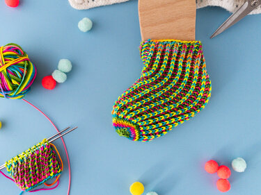 Mehrfarbige gestrickte Babysocke auf Holzblock mit Wollknäuel und Stricknadeln auf blauem Hintergrund
