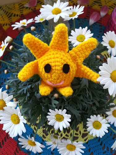 Gelbes gehäkeltes Amigurumi in Sonnenform mit lächelndem Gesicht, umgeben von weißen Gänseblümchen.