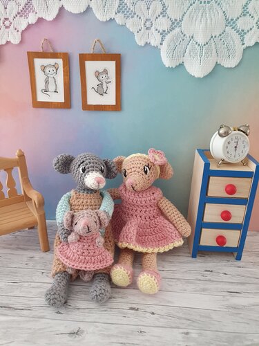 Zwei gehäkelte Amigurumi-Mäuse in Pastellkleidern, eine hält ein kleines Mäusebaby; Holzmöbel und Wecker vor pastellfarbenem Hintergrund