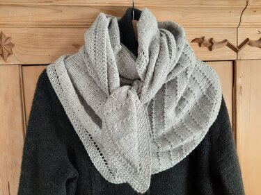 Hellgraues gestricktes Dreieckstuch mit Loch- und Strukturmustern, locker über dunkler Kleidung vor Holzhintergrund drapiert.