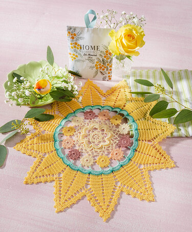 Sternförmiges gelbes Häkeldeckchen mit pastellfarbener Blumenmitte auf rosa Untergrund, daneben gelbe Rose und grüne Zweige