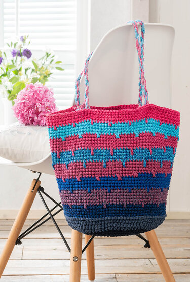 Gehäkelte Shopper-Tasche mit Streifen in Pink, Türkis, Blau und Violett, mit geflochtenen Tragegriffen an einem Stuhl.