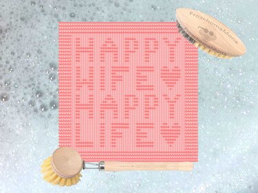 Korallfarbener gestrickter Topflappen mit Spruch ‚HAPPY WIFE HAPPY LIFE‘ und Herzmotiven, arrangiert mit Holzbürsten auf seifigem Hintergrund