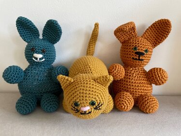 Drei gehäkelte Amigurumi-Stofftiere: eine gelbe Katze und zwei Hasen in Petrol und Rost auf hellem Untergrund.