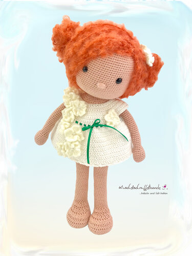 Gehäkelte Amigurumi-Puppe mit rotem lockigem Haar, cremeweißem Kleid mit Blumenapplikationen und grünem Band vor hellem Hintergrund