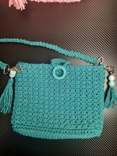 Türkise gehäkelte Umhängetasche mit geflochtenem Riemen, Quasten, Perlen und ringförmigem Verschluss auf dunklem Hintergrund.