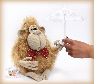 Crocheted shaggy light-brown amigurumi monkey wearing a red polka-dot bow tie, pictured with a small white lace umbrella.
