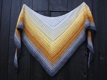 Gehäkeltes Dreieckstuch mit Verlaufsgarn in Creme, Gelb und Grau vor dunkler Holzwand