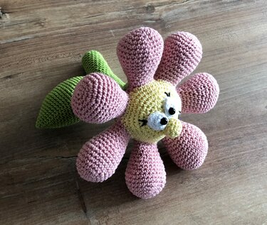 Gehäkelte Blumenrassel mit rosa Blüten, gelbem Gesicht und grünem Blatt auf Holzuntergrund