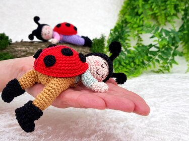 Hand hält gehäkelten, schlafenden Amigurumi-Marienkäfer; weitere Marienkäfer im Hintergrund.
