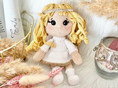 Gehäkeltes Amigurumi-Engelchen mit blonden Locken, Heiligenschein, weißen Flügeln und weißem Kleid mit goldfarbener Borte auf weißem Kunstfell.