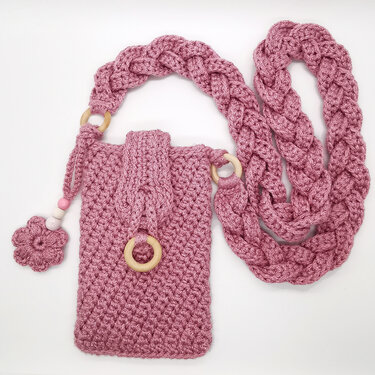 Pink crocheted phone pouch with braided strap, wooden rings and a small flower charm on a white background.